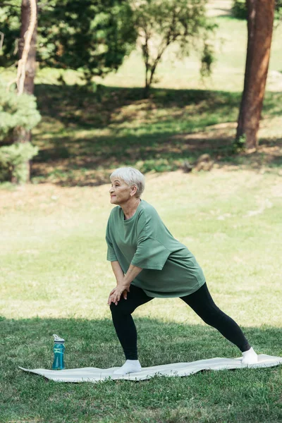 Full length of senior woman with grey hair smiling and doing lunges on fitness mat in park — Foto stock