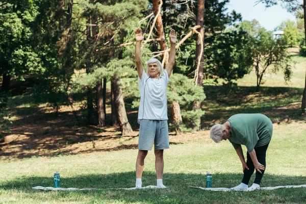 Full length of senior man with grey hair working out on fitness mat near wife in park — Stock Photo