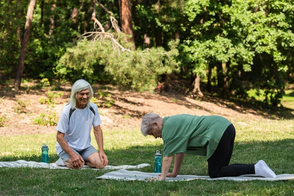 Full length of cheerful senior couple in sportswear exercising on fitness mats in green park — Photo de stock
