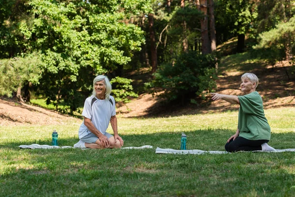 Full length of senior woman pointing with finger near happy husband on fitness mat in green park — Stockfoto