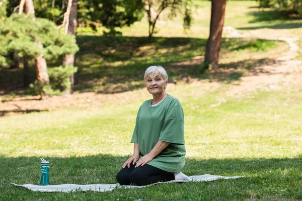 Full length of senior woman with grey hair sitting on fitness mat in park — Stock Photo