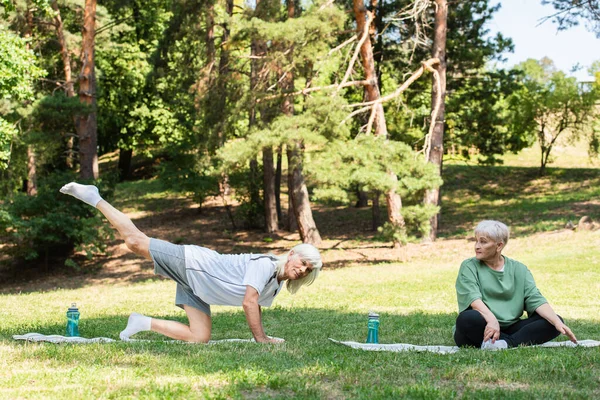 Full length of senior woman in sportswear looking at husband exercising on fitness mat in green park — Fotografia de Stock