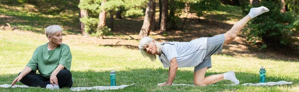 Full length of senior woman in sportswear looking at husband exercising on fitness mat in green park, banner — Stockfoto