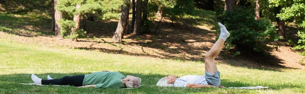 Полная длина пожилой пары в спортивной одежде, работающей на фитнес-ковриках в зеленом парке, баннер — стоковое фото