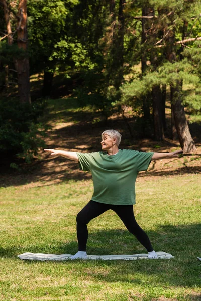 Full length of senior woman with grey hair smiling and working out on fitness mat in park — Stockfoto