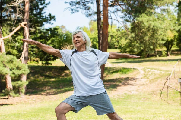Весёлый пожилой мужчина с седыми волосами, улыбающийся и тренирующийся с протянутыми руками в парке — стоковое фото