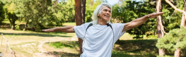 Веселый пожилой мужчина с седыми волосами, улыбающийся и тренирующийся с протянутыми руками в парке, баннер — стоковое фото