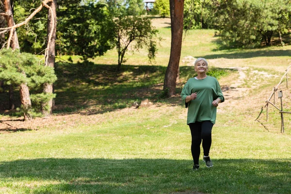Full length of senior woman with grey hair smiling and running on grass in park — Stock Photo
