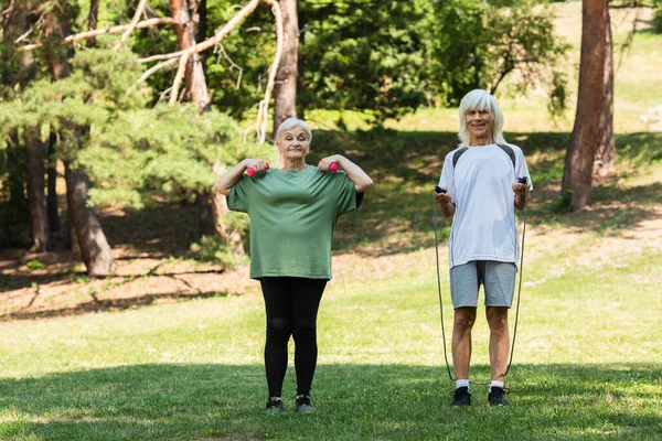 Повна довжина веселої старшої пари в спортивному одязі вправи зі спортивним обладнанням в зеленому парку — стокове фото