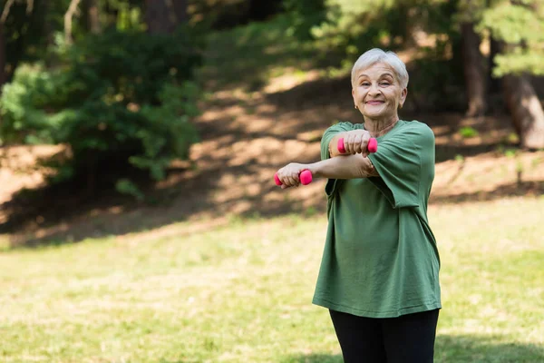 Happy senior woman with grey hair smiling and exercising with dumbbells in park — Stock Photo