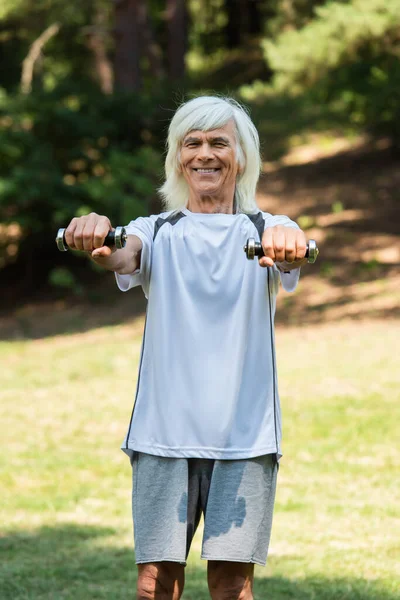 Щасливий старший чоловік у спортивному одязі вправляється з гантелями в зеленому парку — стокове фото