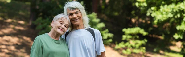 Весела старша пара в спортивному одязі обіймається в зеленому парку, банер — стокове фото