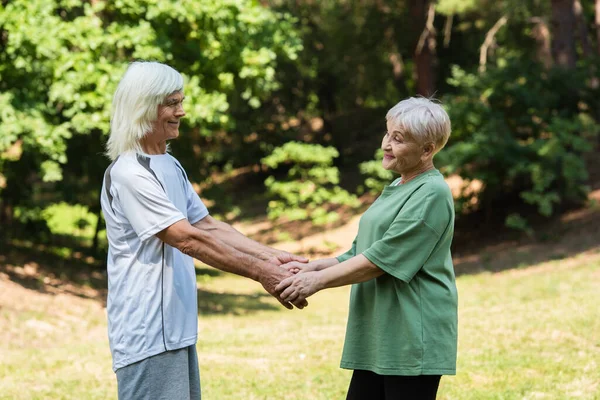Веселая старшая пара в спортивной одежде, держащаяся за руки в зеленом парке — стоковое фото