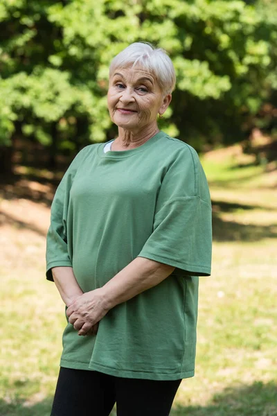 Cheerful and retied woman with grey hair standing in green t-shirt in park — Photo de stock