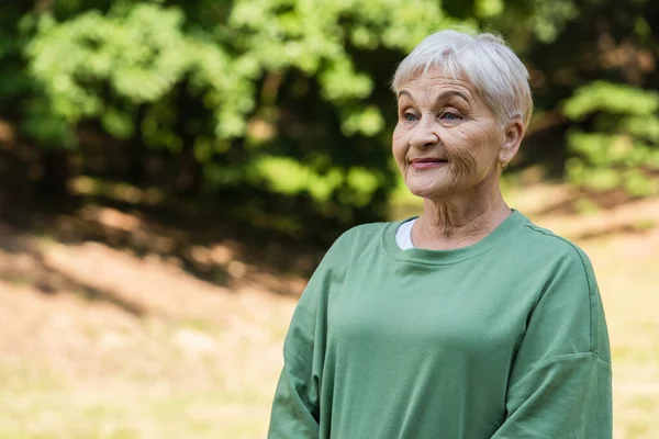 Happy and retied woman with grey hair standing in green t-shirt in park - foto de stock