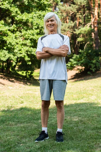 Full length of happy senior man in sportswear standing with crossed arms in green park — Stockfoto