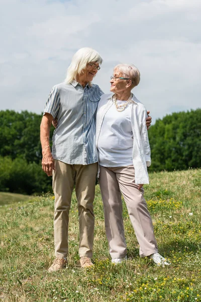 Повна довжина щасливого старшого чоловіка і дружини з сірим волоссям, що стоїть і обіймається на зеленому пагорбі влітку — стокове фото