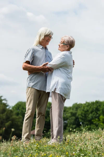 Full length of happy senior husband and wife with grey hair hugging on green hill in summer — Fotografia de Stock