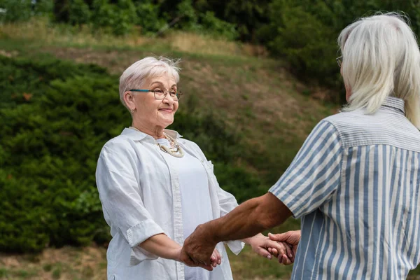 Happy senior woman in glasses holding hands with husband in striped shirt in park — Stockfoto