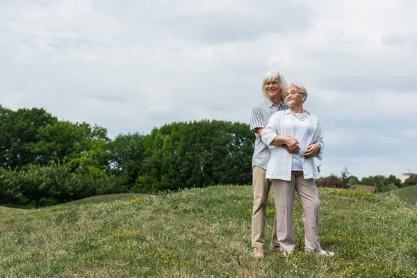 Cheerful senior man in glasses hugging wife with grey hair and standing on green hill — Stockfoto