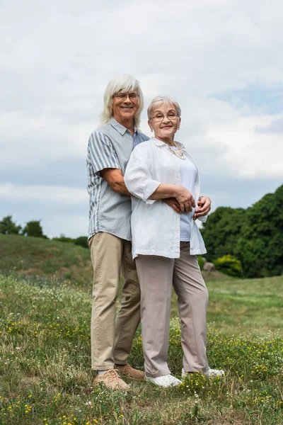 Счастливый пожилой мужчина в очках обнимает счастливую жену с седыми волосами и стоит на зеленом холме — стоковое фото