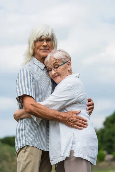 Happy senior husband and wife with grey hair embracing against cloudy sky — Fotografia de Stock