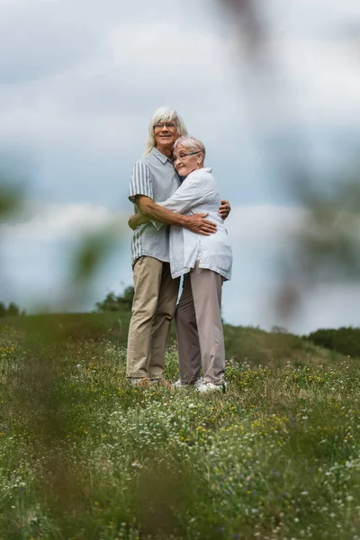 Happy senior husband and wife with grey hair hugging on green hill with blurred foreground — Stockfoto