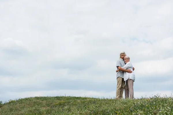 Happy senior husband and wife with grey hair hugging and standing on green hill — Stock Photo