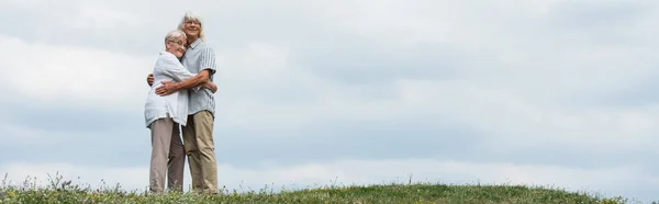 Щасливий старший чоловік і дружина з сірим волоссям обіймаються і стоять на зеленому пагорбі, банер — стокове фото