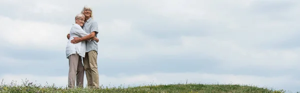 Happy senior husband and wife with grey hair hugging and standing on green hill, banner — Foto stock