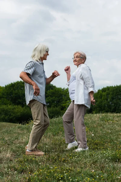 Full length of happy retired couple in glasses dancing on green hill — Fotografia de Stock