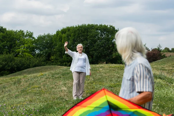 Happy senior woman in casual clothes waving hand at husband with kite on green hill — Stockfoto