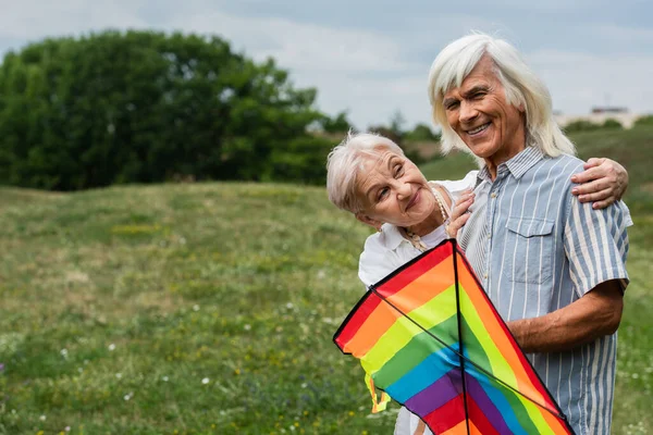 Happy senior woman in casual clothes hugging husband with kite on green hill — Stock Photo