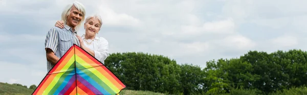 Happy senior woman in casual clothes hugging husband in glasses standing with kite, banner — Stock Photo