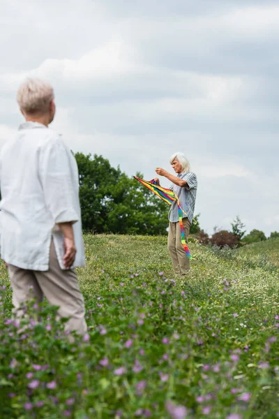 Старший мужчина в повседневной одежде держит воздушного змея и стоит на зеленом холме рядом с размытой женой — стоковое фото