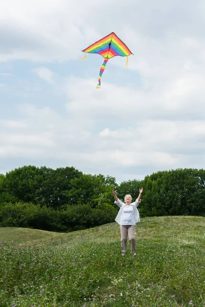Full length of senior woman in casual clothes looking at flying kite in cloudy sky — Photo de stock