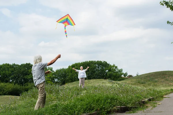 Happy senior woman in casual clothes looking at husband playing with kite on green hill — Fotografia de Stock