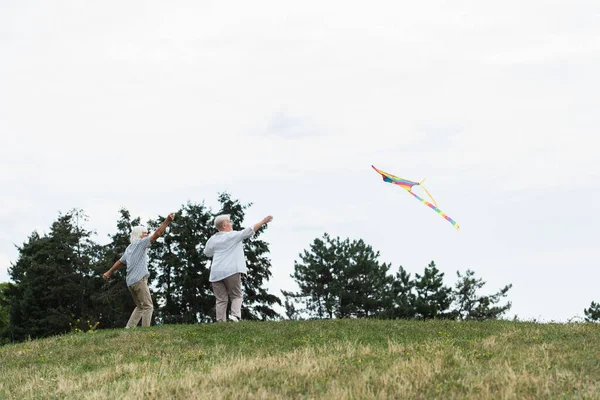 Senior couple in casual clothes playing with kite on green hill — Stockfoto