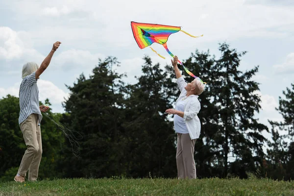 Happy senior woman in casual clothes playing with kite near husband on green hill — Foto stock
