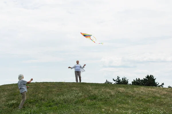 Cheerful senior woman in casual clothes looking at kite near husband on green hill — Stock Photo