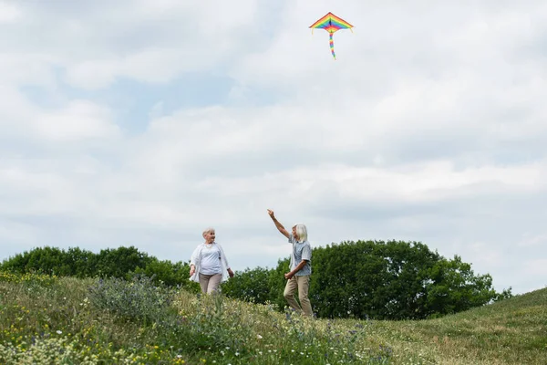 Happy senior couple in casual clothes playing with kite on green hill — Stockfoto