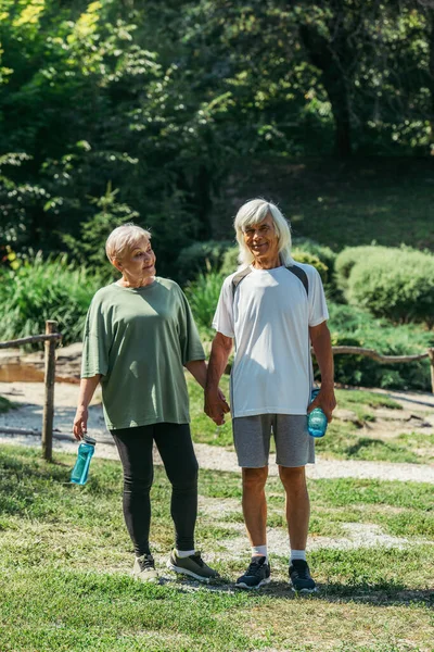 Полная длина счастливой пары на пенсии в спортивной одежде держа спортивные бутылки и руки друг друга в парке — стоковое фото