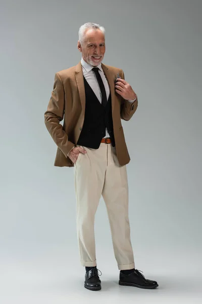 Full length of joyful middle aged groom in suit with floral boutonniere standing with hand in pocket on grey — Foto stock