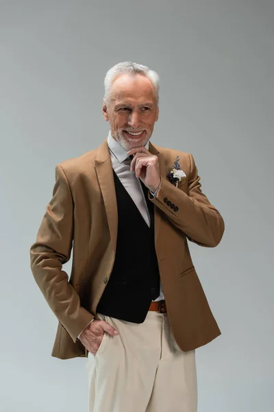 Happy middle aged groom in suit with floral boutonniere posing with hand in pocket isolated on grey — Foto stock