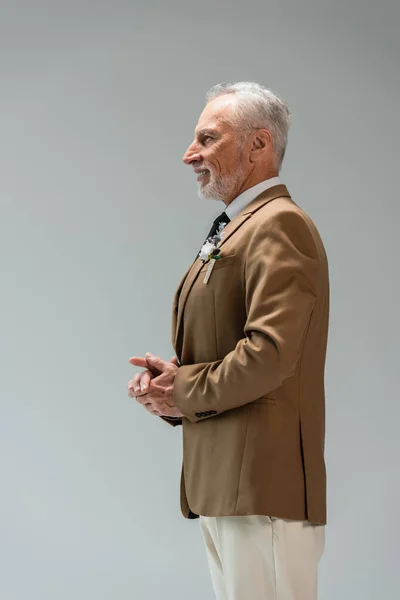 Side view of happy middle aged groom in suit with floral boutonniere isolated on grey — Stockfoto