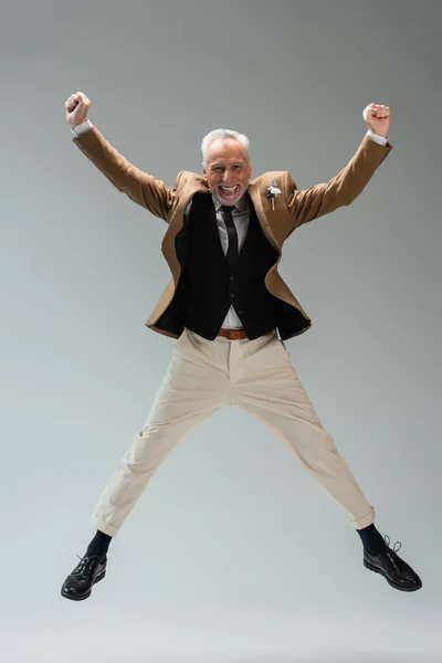 Full length of happy middle aged groom in suit with floral boutonniere jumping on grey - foto de stock