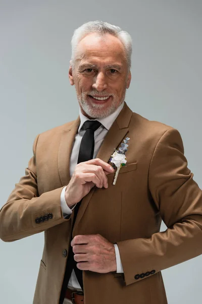 Happy middle aged groom in beige blazer adjusting floral boutonniere isolated on grey — Stockfoto