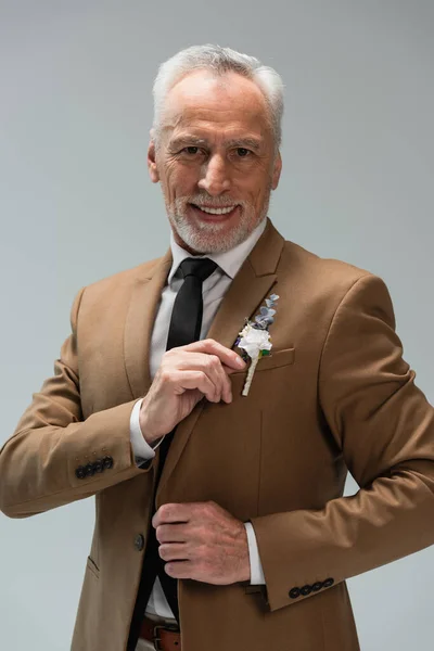 Happy middle aged groom in beige blazer adjusting floral boutonniere isolated on grey — Foto stock