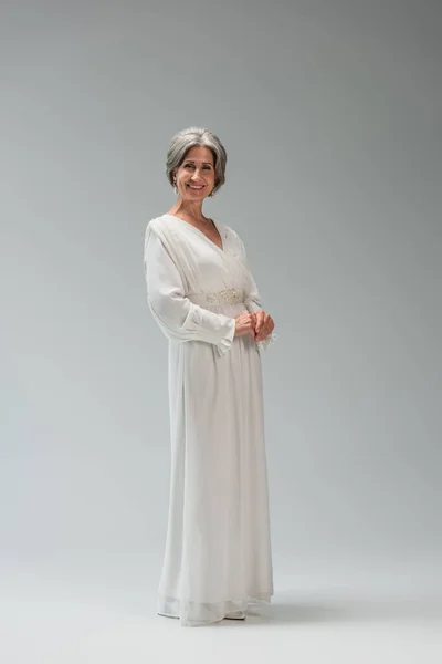 Повна довжина щасливої нареченої середнього віку в білій весільній сукні, що стоїть на сірому — Stock Photo