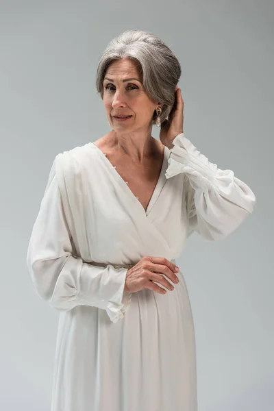 Middle aged bride in white wedding dress adjusting grey hair isolated on grey — Stockfoto